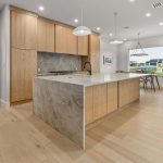 Modern kitchen with light wood cabinets, a large marble island with a built-in sink, pendant lights, light wood floors, and an adjacent dining area with large windows showing a view outside.