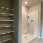 A bathroom with a walk-in shower featuring white panels and built-in shelves, an open doorway leading to a carpeted walk-in closet, and a separate nook with five white wall-mounted shelves.