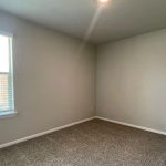 Empty room with beige carpet, light gray walls, a window with white blinds letting in daylight, and a ceiling light. There are electrical outlets visible on the walls.