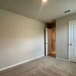 A small empty room with beige walls, carpeted floor, a closed white door, an open door leading to a hallway, and a ceiling vent. Natural light enters from an unseen window.