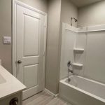 A modern bathroom with a white sink and countertop on the left, a white door in the center, and a white bathtub with built-in shelves and shower on the right. Light-colored walls and floor tiles complete the look.