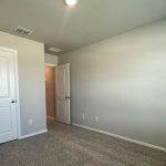 A small empty room with beige walls, a closed white door on the left, an open white door on the right, and light brown carpeted floor. A ceiling light is on, and the room is unfurnished.
