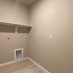 Empty laundry room with beige walls, a single white shelf, electrical outlets, water hookups, and a vent opening near the bottom. Light wood flooring covers the floor. The space appears clean and unfurnished.