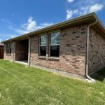 Single-story brick house with two large windows facing a green, well-maintained backyard enclosed by a wooden fence, under a sunny blue sky with scattered clouds.