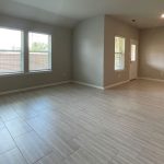 Spacious, empty living area with large windows, light-colored tile flooring, beige walls, and an open doorway leading to another room. A door and additional window are visible in the adjacent area.