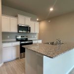 Modern kitchen with white cabinets, stainless steel appliances, a granite countertop island with sink, light wood-style flooring, and neutral-colored walls under recessed lighting.
