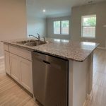 A modern kitchen with a granite island that includes a sink and dishwasher, opening to a bright living area with large windows, light wood tile flooring, and a white door leading outside.