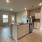 Modern kitchen with white cabinets, granite countertops, stainless steel appliances, and an island with a sink. The open layout leads to a bright living area with large windows and light-colored tile flooring throughout.