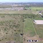Aerial view of a grassy and partly wooded field with scattered trees, several dirt paths, a parking area with a few vehicles, and buildings in the background, surrounded by suburban homes and open land.