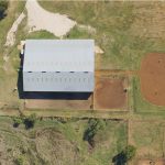 Aerial view of a large rectangular barn or arena, two round horse arenas with horses, several parked vehicles on grass, and fenced grassy areas in a rural landscape.