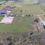 Aerial view of a large open field with green grass, a rectangular building with several parked vehicles nearby, a dirt road, some fenced areas, and a cluster of buildings in the upper left corner. Trees and rural landscape surround the area.
