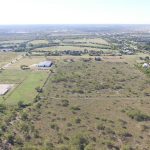 Aerial view of a large rural landscape with open fields, scattered trees, and a few buildings. Residential houses are visible to the right, and farmland stretches out to the horizon under a clear sky.
