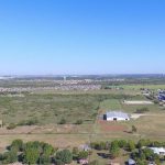 Aerial view of a rural landscape with open fields, scattered trees, a few buildings, and a large white-roofed structure under a clear blue sky. Distant neighborhoods and city skyline are visible on the horizon.