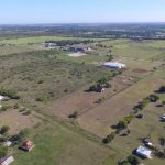 Aerial view of a rural landscape featuring open fields, scattered houses, farm buildings, roads, and patches of trees under a clear blue sky. Residential areas and farmland stretch toward the horizon.
