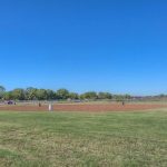 A baseball diamond with a dirt infield and grassy outfield sits in an open park under a clear blue sky, surrounded by trees and a distant fence.