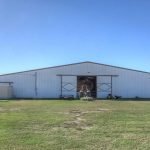 A large white metal barn with open double doors sits on a grassy field under a clear blue sky. Various equipment and items are visible near the entrance and sides of the building.