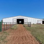 A large white metal barn with open doors stands at the end of a dirt path, flanked by fenced areas and surrounded by grass under a clear blue sky.