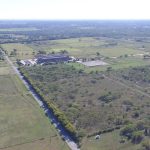 Aerial view of a rural area with open fields, scattered trees, roads, and a few buildings under a clear sky. The landscape extends into the distance with greenery and farmland.