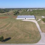 Aerial view of a large barn or stable surrounded by vast grassy fields, dirt road leading up to the building, with a few vehicles and scattered trees, under a clear blue sky.