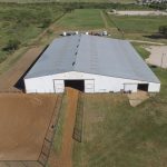 Aerial view of a large white barn with a metal roof next to a fenced dirt riding arena, surrounded by grassy fields and a few scattered vehicles.
