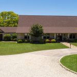 A large, single-story house with a brown roof, stone and brick exterior, surrounded by a well-manicured lawn, trees, and a circular gravel driveway under a clear blue sky.