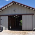 A large metal barn with an open sliding door, revealing the interior. The barn sits on a gravel area under a blue sky with wispy clouds. Some metal equipment and a hose are visible outside.