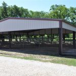 A large, open-sided metal barn with a peaked roof and fenced enclosures, situated on gravel and surrounded by grass and trees.