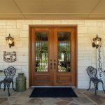 A double wooden front door with glass panels is flanked by two black lanterns, metal chairs, and small tables on a stone patio. A vertical "WELCOME" sign and a green jug sit near the entrance.