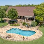 Aerial view of a large house with a sloped roof, surrounded by trees and a spacious lawn. In the backyard, there is a stone patio with a swimming pool and lounge chairs arranged around it.