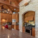 Rustic kitchen with stone and wood details, stainless steel appliances, wooden cabinets, granite countertops, and a mounted longhorn above the stove. Exposed beams and archways add to the warm, country-style atmosphere.