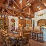 Spacious rustic kitchen and dining area with wooden beams, vaulted ceiling, stone walls, large wooden table with chairs, bar counter with stools, glass cabinets, and stainless steel appliances. Warm, inviting atmosphere.