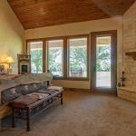 A rustic bedroom with a wooden bed, star decorations on the wall, large windows with blinds, and a stone fireplace with a wooden mantel. Warm lighting and a view of trees outside complete the cozy atmosphere.