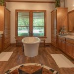 Spacious bathroom with large freestanding tub centered between two wooden vanities with sinks, marble countertops, mirrors, and windows letting in natural light. Wooden floors and a round rug complete the warm decor.