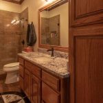 A warm-toned bathroom with wood cabinets, a marble countertop, a large mirror, a toilet, and a walk-in shower with tan tiles and a glass door. Towels hang on the wall, and the floor features a patterned rug.