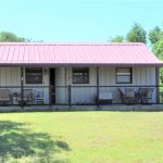 A small, single-story house with a red metal roof, front porch, and fenced yard, surrounded by trees. There are chairs on the porch, a satellite dish, and a small shed to the right. The lawn is green and sunlit.