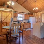 A cozy kitchen and dining area with wood-paneled walls and ceiling, a round dining table with chairs, a kitchen island with stools, barn-style double doors, and wooden cabinets. Warm lighting creates an inviting atmosphere.
