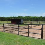A fenced paddock area with black metal gates and a small shelter stands on grassy land, surrounded by trees and open fields under a blue sky.