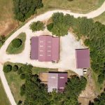 Aerial view of a rural property with several red-roofed buildings surrounded by trees, grassy areas, and a winding gravel road. The buildings are arranged in a roughly rectangular layout.