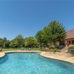 A backyard with a curved swimming pool surrounded by a stone patio, outdoor seating, trees, and greenery under a clear blue sky, next to a large house with a shaded porch.