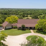 Aerial view of a large, single-story house with a red roof, surrounded by green lawns, trees, and a circular driveway. A white SUV is parked outside, and open fields and forests are visible in the background.