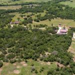 Aerial view of a large rural property with several buildings surrounded by green fields, trees, winding dirt roads, and patches of forested land. The landscape appears hilly and natural.