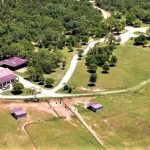 Aerial view of a ranch with several buildings, fenced paddocks, a winding road, scattered trees, and open grassy areas, surrounded by a dense wooded area.