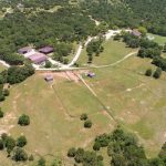 Aerial view of a large rural property with several buildings, open fields, scattered trees, and dirt roads winding through green landscape. The area appears peaceful and spacious, surrounded by dense wooded areas.