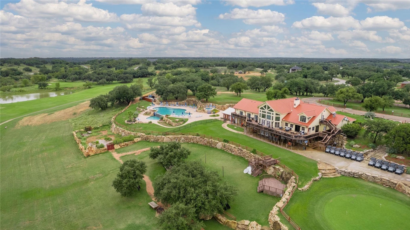 Aerial view of a country club with a large swimming pool, clubhouse with red roof, golf carts, putting green, stone fences, trees, and open grassy landscape under a partly cloudy sky.