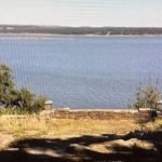 A view of a large, calm lake with a stone wall and a sign reading "Hiking Trail" in the foreground. Trees and grass frame the scene, and distant shoreline is visible under a clear sky.