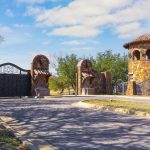 A grand stone entrance with an ornate iron gate and two-story turret, surrounded by trees and blue sky with scattered clouds. The roadway curves toward the gated entrance.