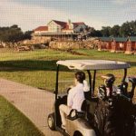 A person driving a golf cart with golf clubs on a paved path, passing by green grass and heading toward a large clubhouse and several cabin-style buildings in the background.