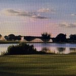 A serene lake scene at sunset with fountains spraying water, trees silhouetted in the background, and a lush green lawn in the foreground under a partly cloudy sky.