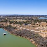 Aerial view of a rural lakeside area with a dock extending into greenish water, winding dirt roads, scattered trees, and a few buildings under a clear blue sky.