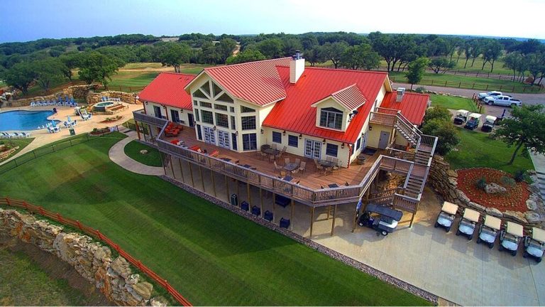 A large, two-story building with a red roof and wide balcony sits on a manicured lawn, next to a driveway lined with golf carts; a swimming pool and trees are visible in the background.